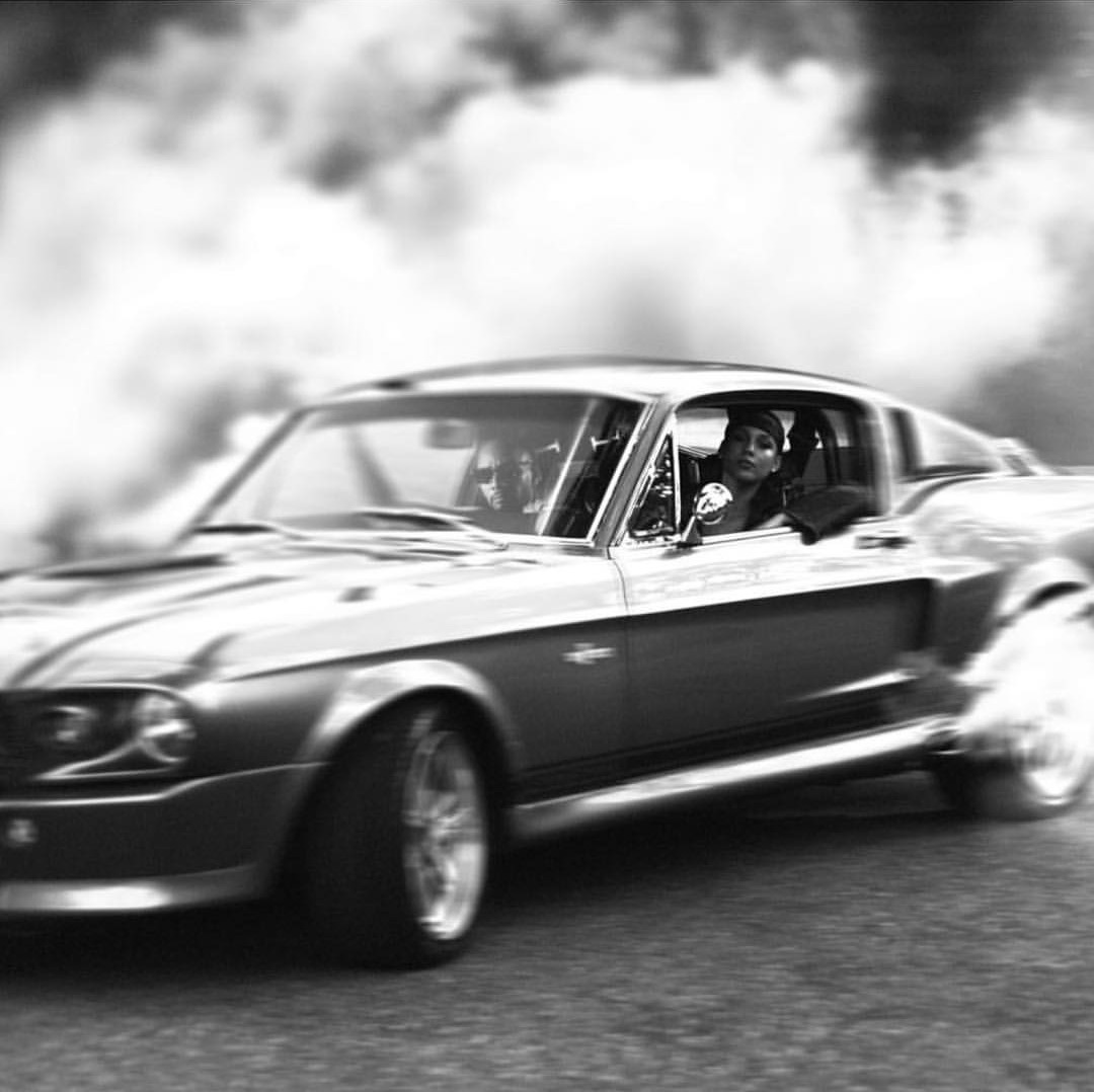 A Shelby Mustang fastback in motion, shot in black and white, smoke pouring off the rear wheels and a woman watching from the passenger window